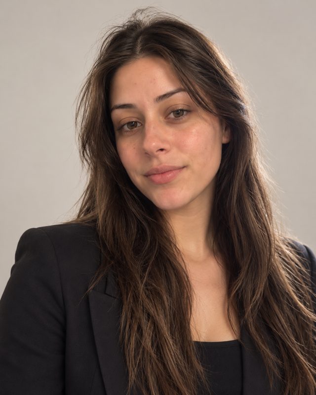 Example of the Studio headshot look: a woman with long dark hair in a black blazer on a light-grey studio backdrop